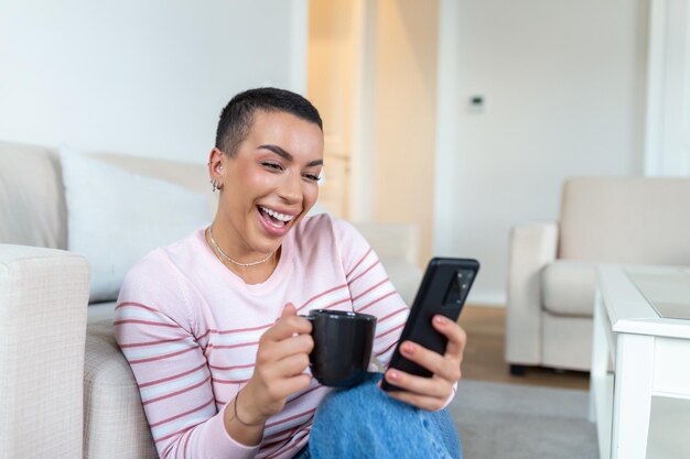 Una mujer joven en un suéter con una taza en las manos mira el teléfono mientras está sentada en el sofá