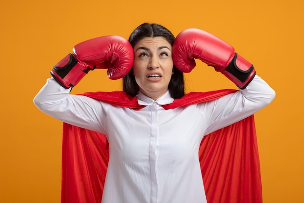 Mujer joven superhéroe dolorida con guantes de box golpeándose a sí misma en la cabeza mirando hacia arriba aislado en la pared naranja