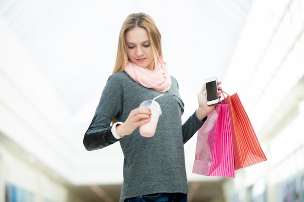Mujer joven en el supermercado comprobación de la celebración de bolsas de la compra, teléfono móvil y una bebida