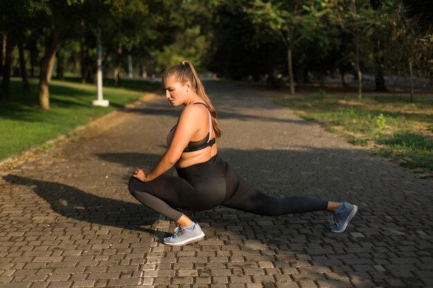 Mujer joven de talla grande con top deportivo y calzas cuidadosamente haciendo ejercicios deportivos mientras pasa tiempo en el acogedor parque de la ciudad