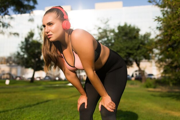 Mujer joven de talla grande con top deportivo y polainas con auriculares rojos mirando cansadamente a un lado mientras se apoya en las rodillas en el parque de la ciudad