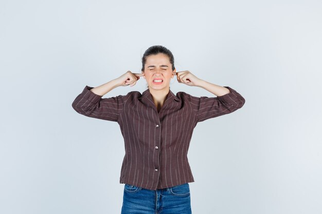 Mujer joven tapando los oídos con los dedos índices en camisa a rayas, jeans y mirando molesto, vista frontal.