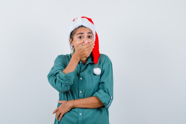 Mujer joven tapándose la boca con la mano, vistiendo un gorro de Papá Noel y mirando consternado. vista frontal.