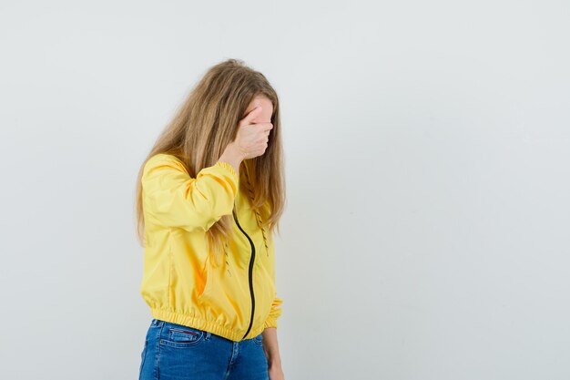 Mujer joven tapándose los ojos con la mano en chaqueta de bombardero amarilla y jean azul y mirando tímida. vista frontal.