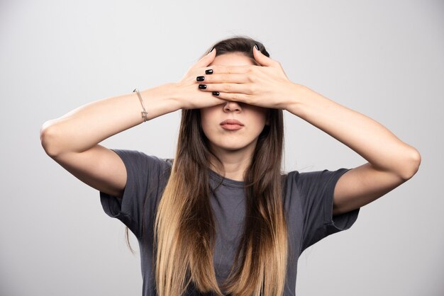 Mujer joven tapándose los ojos con las manos sobre un fondo gris.