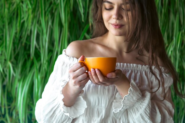 Una mujer joven con una taza de café en el fondo con plantas verdes.