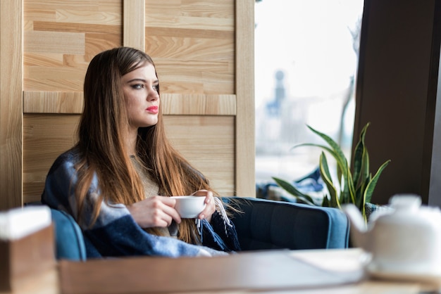 Mujer joven con taza de café mirando a través de la ventana