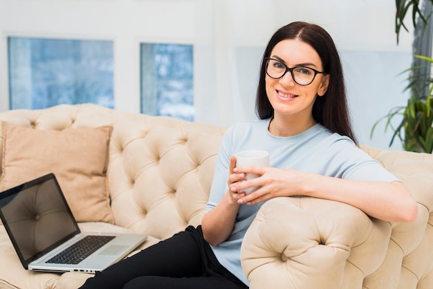 Mujer joven con la taza de café que se sienta en el sofá