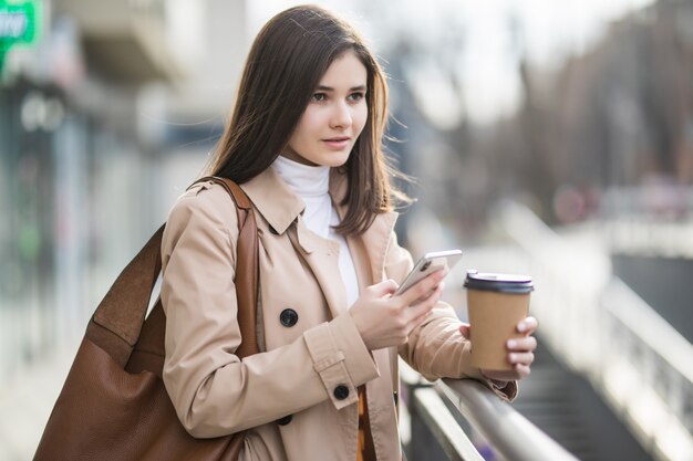 Mujer joven con taza de café por teléfono en la ciudad