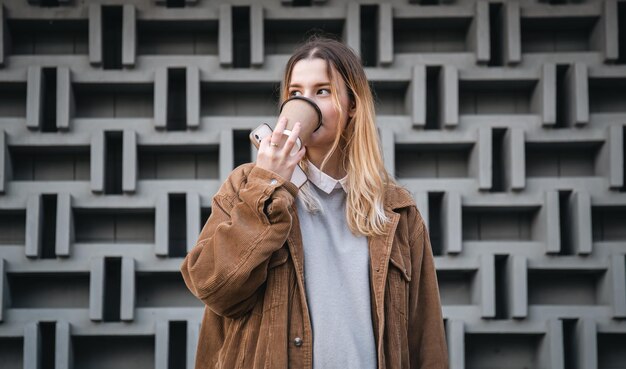 Una mujer joven con una taza de café y un teléfono inteligente en el fondo de una pared.