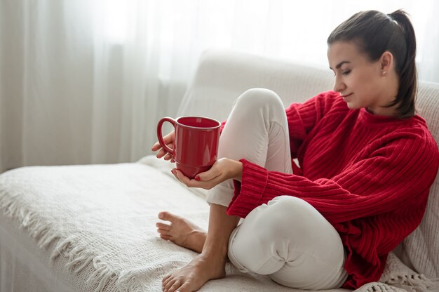 Una mujer joven con una taza roja de bebida caliente en un acogedor suéter rojo está descansando en el sofá en casa copia espacio.