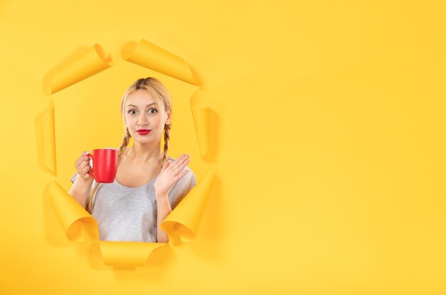 Mujer joven con taza de té en el fondo de papel amarillo rasgado comercial publicidad facial