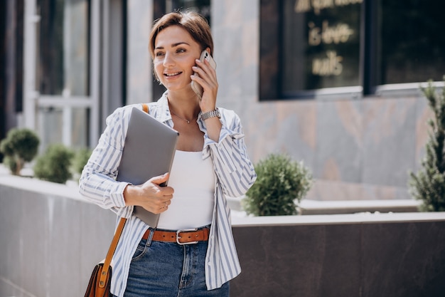 Mujer joven con teléfono y computadora caminando fuera de la calle