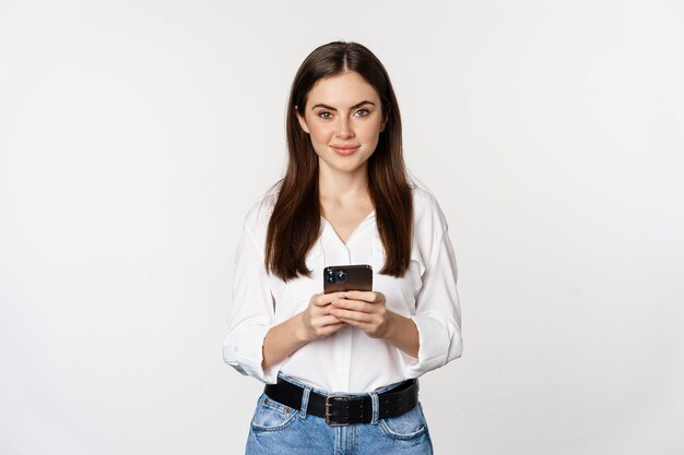 Mujer joven con teléfono inteligente, sonriendo y mirando la cámara, usando una aplicación de teléfono móvil, tecnología celular y concepto de compras en línea, fondo blanco