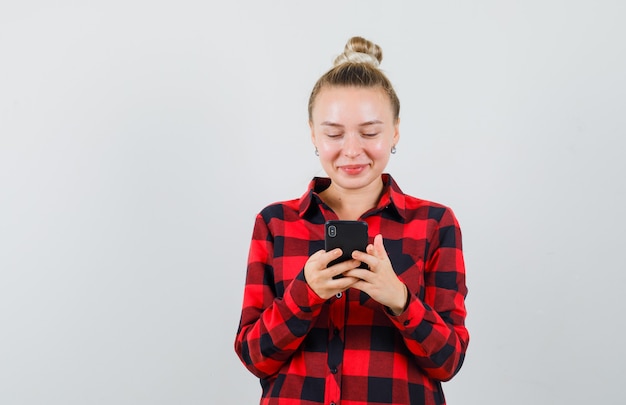 Mujer joven con teléfono móvil en camisa a cuadros y mirando feliz. vista frontal.