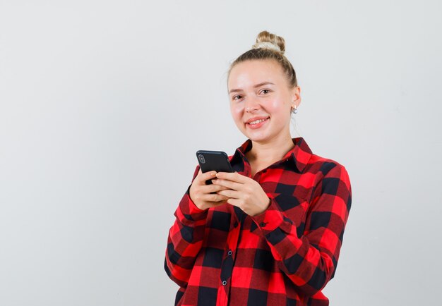 Mujer joven con teléfono móvil en camisa a cuadros y mirando feliz. vista frontal.