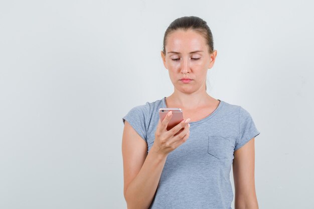 Mujer joven con teléfono móvil en camiseta gris y mirando ocupado, vista frontal.