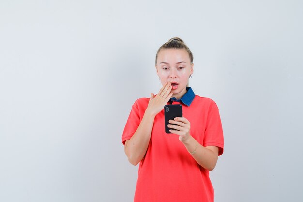 Mujer joven con teléfono móvil en camiseta y mirando sorprendido, vista frontal.