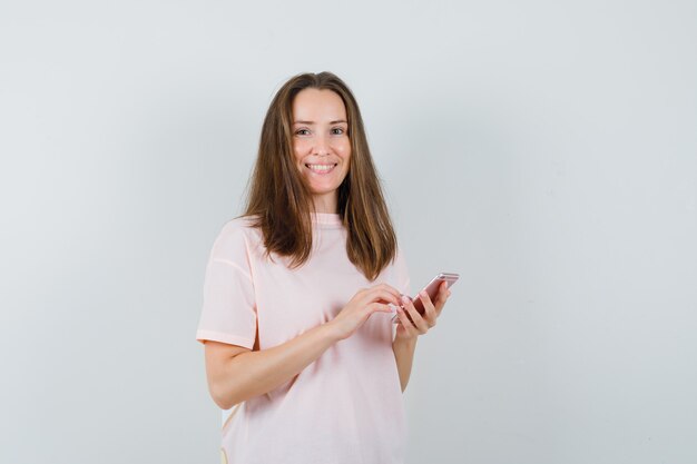 Mujer joven con teléfono móvil en camiseta rosa y mirando contento. vista frontal.