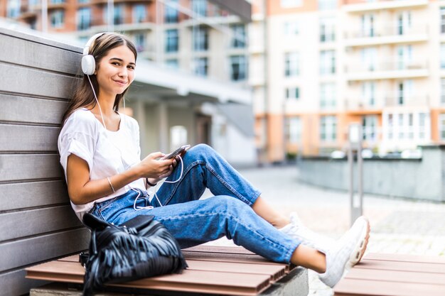 Mujer joven con teléfono móvil escucha música mientras está sentado en el banco en un parque