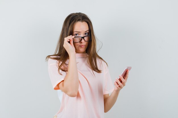 Mujer joven con teléfono móvil, quitándose las gafas en camiseta rosa, vista frontal.