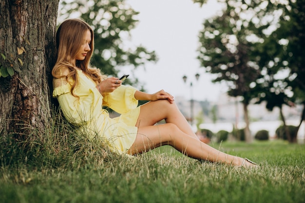 Mujer joven con teléfono y sentado bajo el árbol en el parque