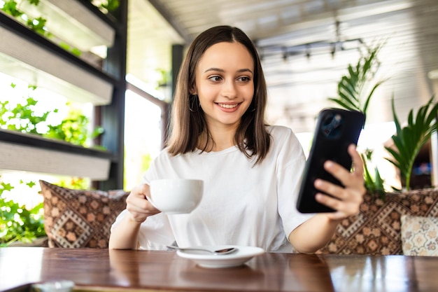 Mujer joven con teléfono sentado fuera del café con bebida caliente