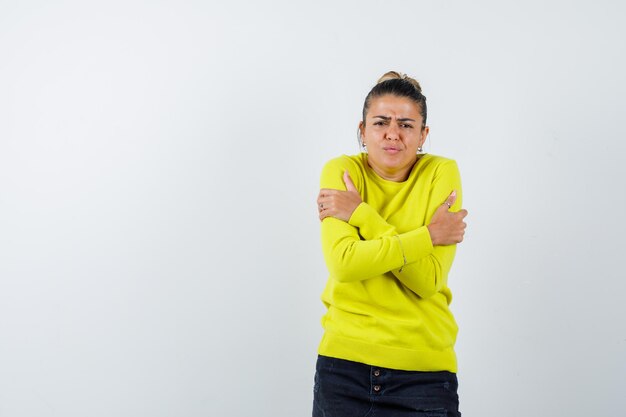 Mujer joven temblando de frío, haciendo muecas en suéter amarillo y pantalón negro y luciendo agobiada