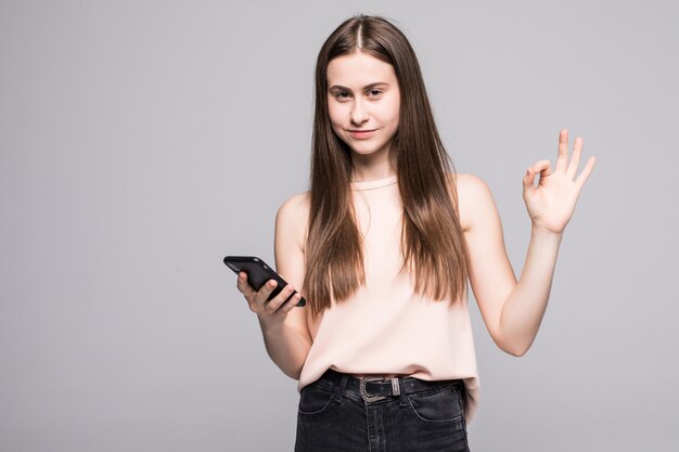 Mujer joven teniendo una conversación hablando por teléfono inteligente sobre pared aislada haciendo aceptar firmar con los dedos