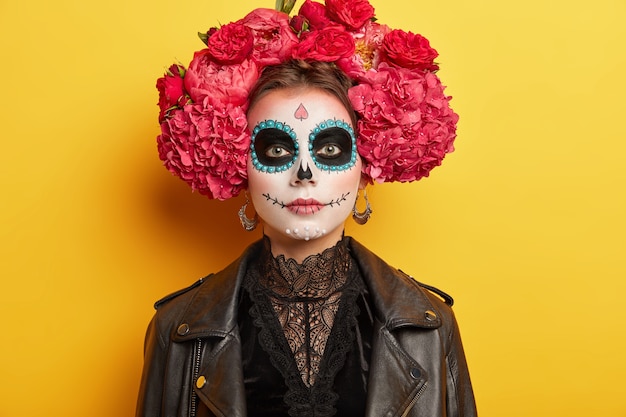 La mujer joven tiene un maquillaje y un disfraz originales, usa una corona de flores rojas, tiene una perspectiva tradicional para las vacaciones mexicanas de dos días, solated en amarillo