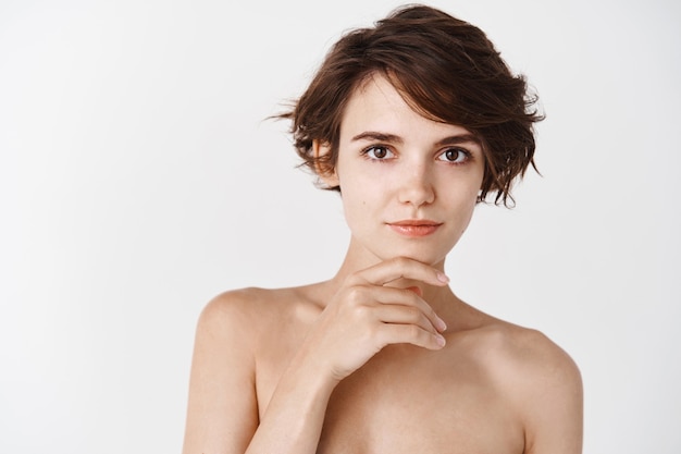 Mujer joven y tierna sin maquillaje, piel limpia hidratada e hidratada, tocando la barbilla y sonriendo de pie desnuda sobre una pared blanca