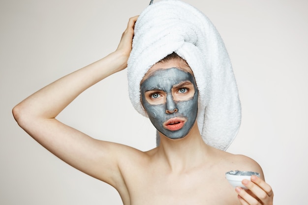 Mujer joven en toalla en la cabeza con máscara facial frunciendo el ceño. Belleza y cuidado de la piel.
