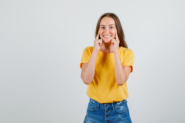 Mujer joven tocando las mejillas con los dedos en camiseta, pantalones cortos y luciendo alegre. vista frontal.