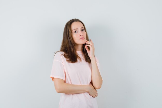 Mujer joven tocando la piel de su rostro en la barbilla en camiseta rosa y mirando tranquila, vista frontal.