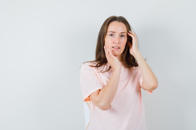 Mujer joven tocando la piel de su rostro en camiseta rosa y luciendo elegante, vista frontal.