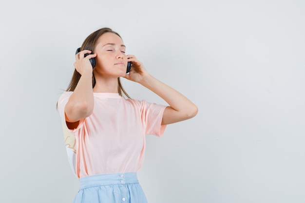 Mujer joven tocando sus auriculares en camiseta, falda y mirando relajado. vista frontal.