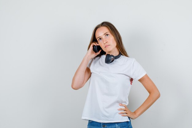 Mujer joven tomados de la mano en los auriculares y la cintura en camiseta blanca, pantalones cortos y mirando pensativo