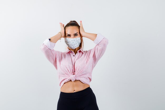 Foto gratuita mujer joven tomados de la mano en la cabeza en camisa, pantalón, máscara médica y mirando olvidadizo, vista frontal.