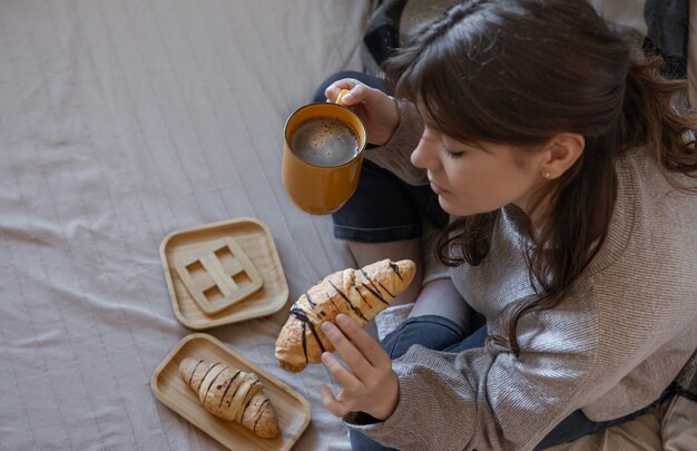 Mujer joven tomando café con un croissant en la cama, vista superior.