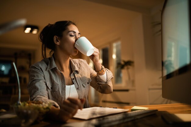 Mujer joven tomando café con los ojos cerrados mientras estudia por la noche en casa