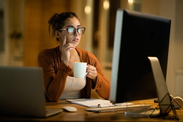 Mujer joven tomando café y pensando mientras trabaja en una PC de escritorio por la noche en casa