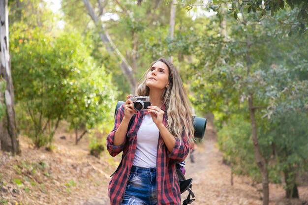 Mujer joven tomando fotos del paisaje con cámara y de pie en el camino forestal. Turista mujer caucásica de pelo largo disparar la naturaleza en el bosque. Turismo de mochilero, aventura y concepto de vacaciones de verano.