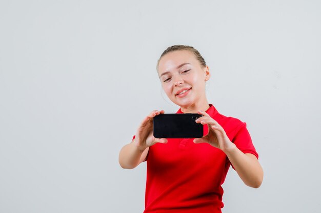 Mujer joven tomando fotos en el teléfono móvil en camiseta roja y mirando alegre