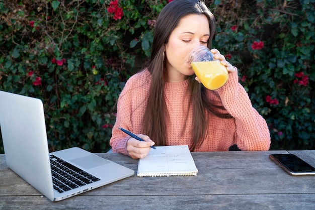 Foto gratuita mujer joven tomando notas al aire libre mientras usa una computadora portátil y bebe jugo de naranja