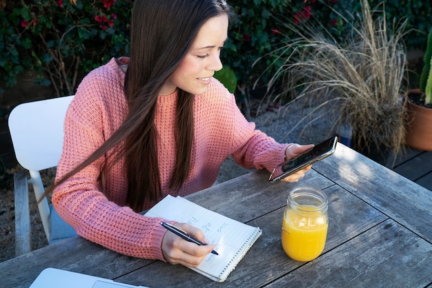 Foto gratuita mujer joven tomando notas al aire libre mientras usa un teléfono inteligente