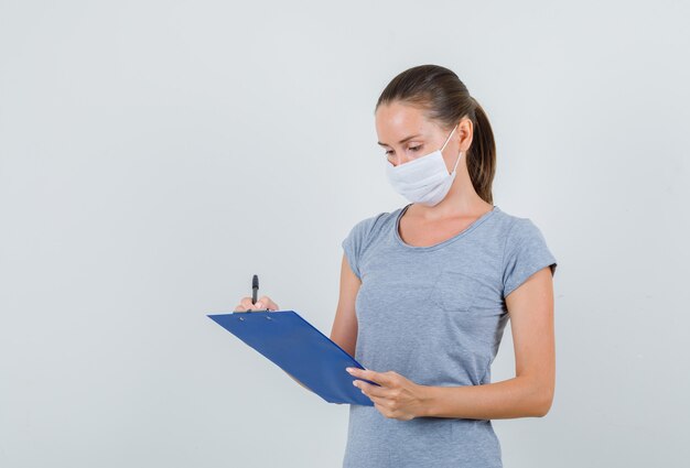 Mujer joven tomando notas en el portapapeles en camiseta gris, máscara y luciendo ocupado. vista frontal.