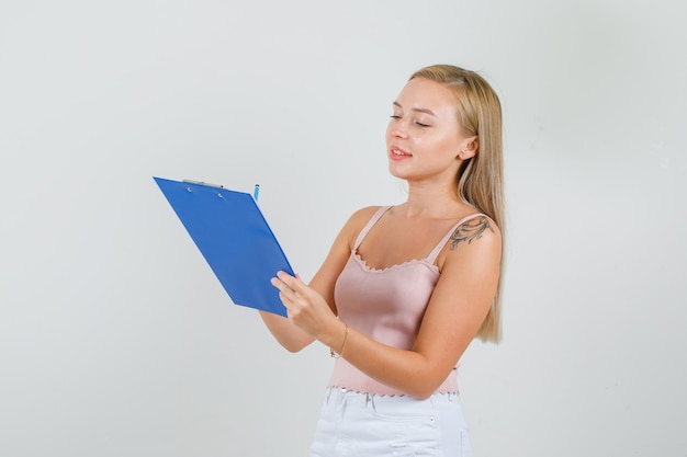 Mujer joven tomando notas en el portapapeles en camiseta, minifalda y mirando ocupado.