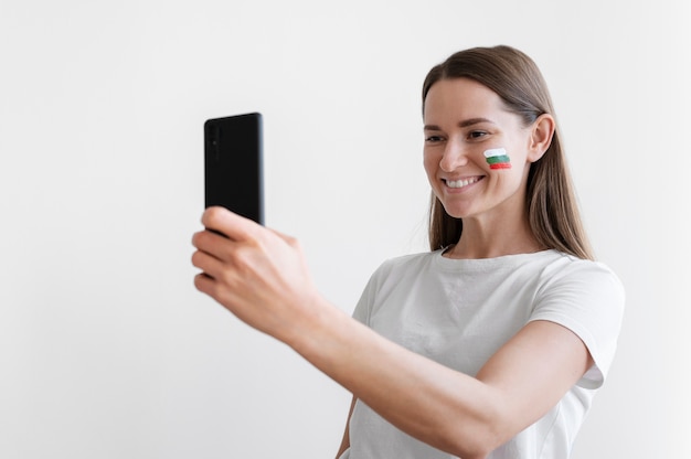 Mujer joven tomando selfie con bandera búlgara pintada en la mejilla