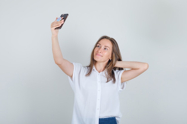 Mujer joven tomando selfie con la mano detrás de la cabeza en camiseta, jeans y mirando alegre