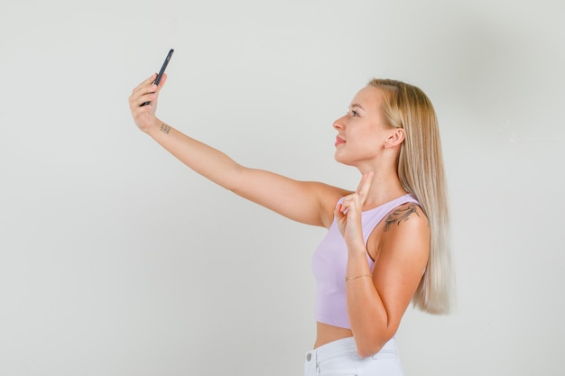 Mujer joven tomando selfie mostrando v-sign en camiseta, minifalda.